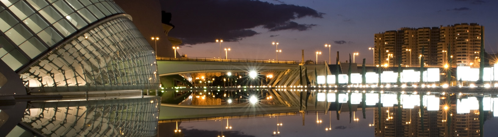 Paisaje urbano nocturno con un edificio moderno de cristal iluminado, un puente encendido y reflejos en agua tranquila bajo un cielo violáceo